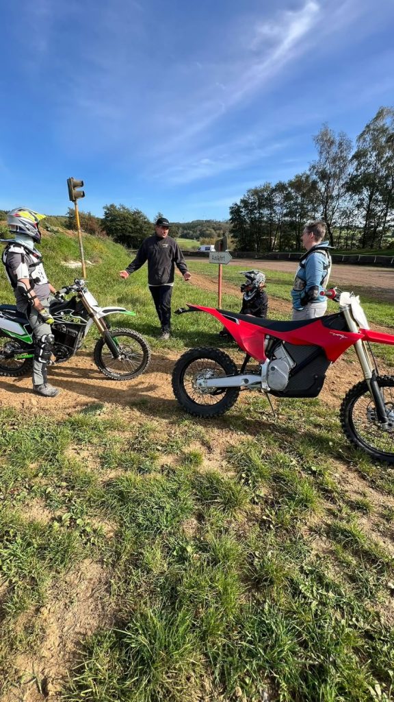 Familienkurs bei Motocross Land: ein Mann in schwarzer Jacke erklärt etwas, um ihn herum zwei Fahrer in Schutzausrüstung, ein Kind mit Helm und ein grün-weißes und ein rotes Motorrad an der Motocross-Strecke