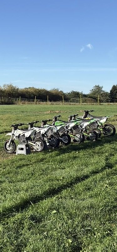 Mehrere kleine grün-weiße Motocross-Motorräder in einer Reihe auf einer Wiese unter blauem Himmel